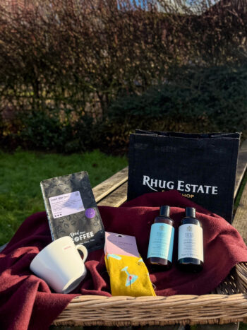 Her hamper - containing, tiered mums club hot chocolate, carried mug, rhug wild beauty shampoo and conditioner and miss sparrow yellow, cocktail motif socks. In a small, black, rhug estate farm shop hessian bag.